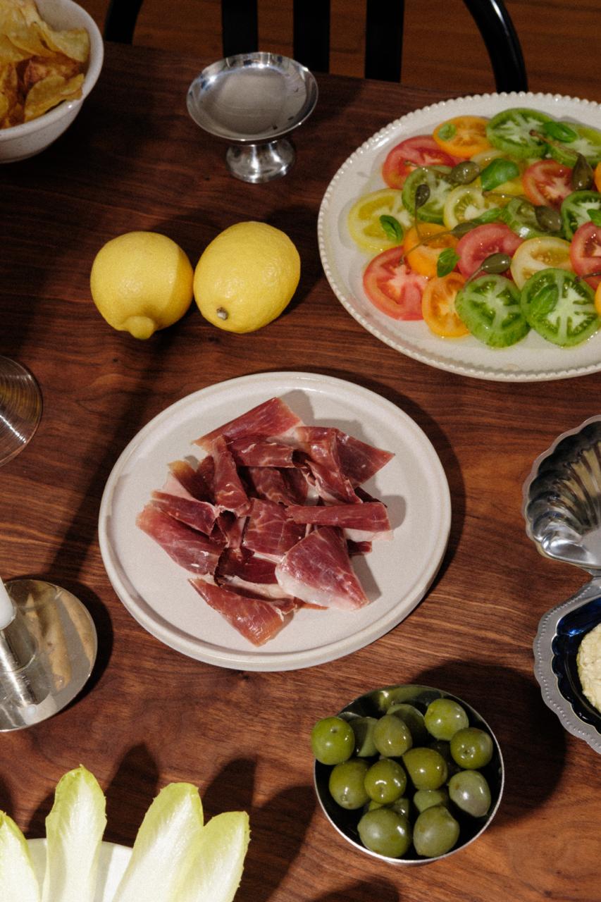 Sur cette table, il y'a plusieurs assiette. Cette scène représente un apéritif.
Au centre, une assiette de Jamón de Cebo de Campo - 50% raza ibérica de la marque LOLO
