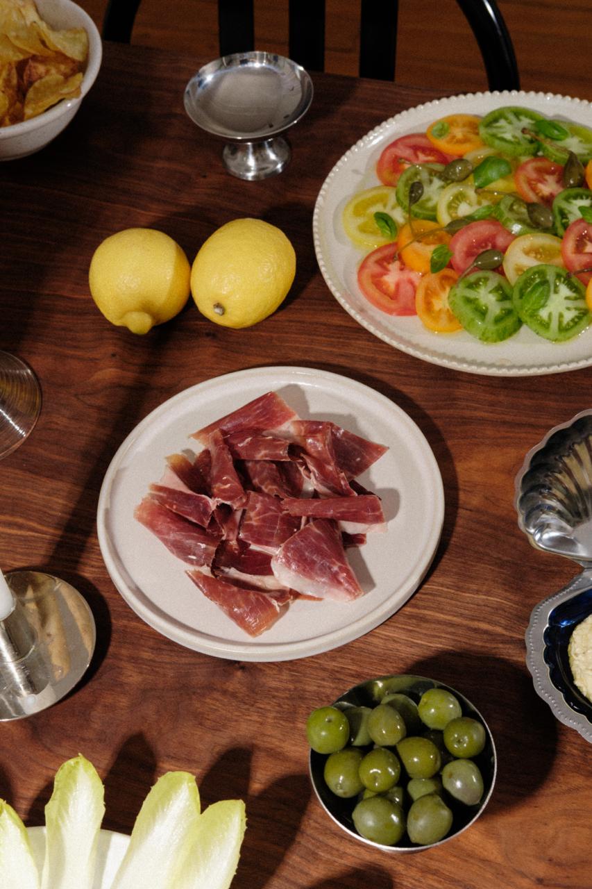 Sur cette table, il y'a plusieurs assiette. Cette scène représente un apéritif. 
Au centre, une assiette de Jamón de Cebo de Campo - 50% raza ibérica de la marque LOLO